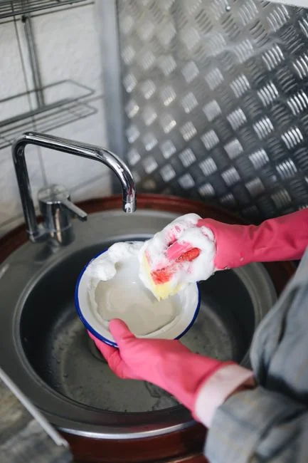 services-04 An anonymous person in yellow latex gloves scrubbing a golden faucet with a sponge in a modern bathroom while cleaning the house